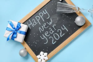 Blackboard With Text HAPPY NEW YEAR 2024, Glass And Christmas Decorations On Blue Background