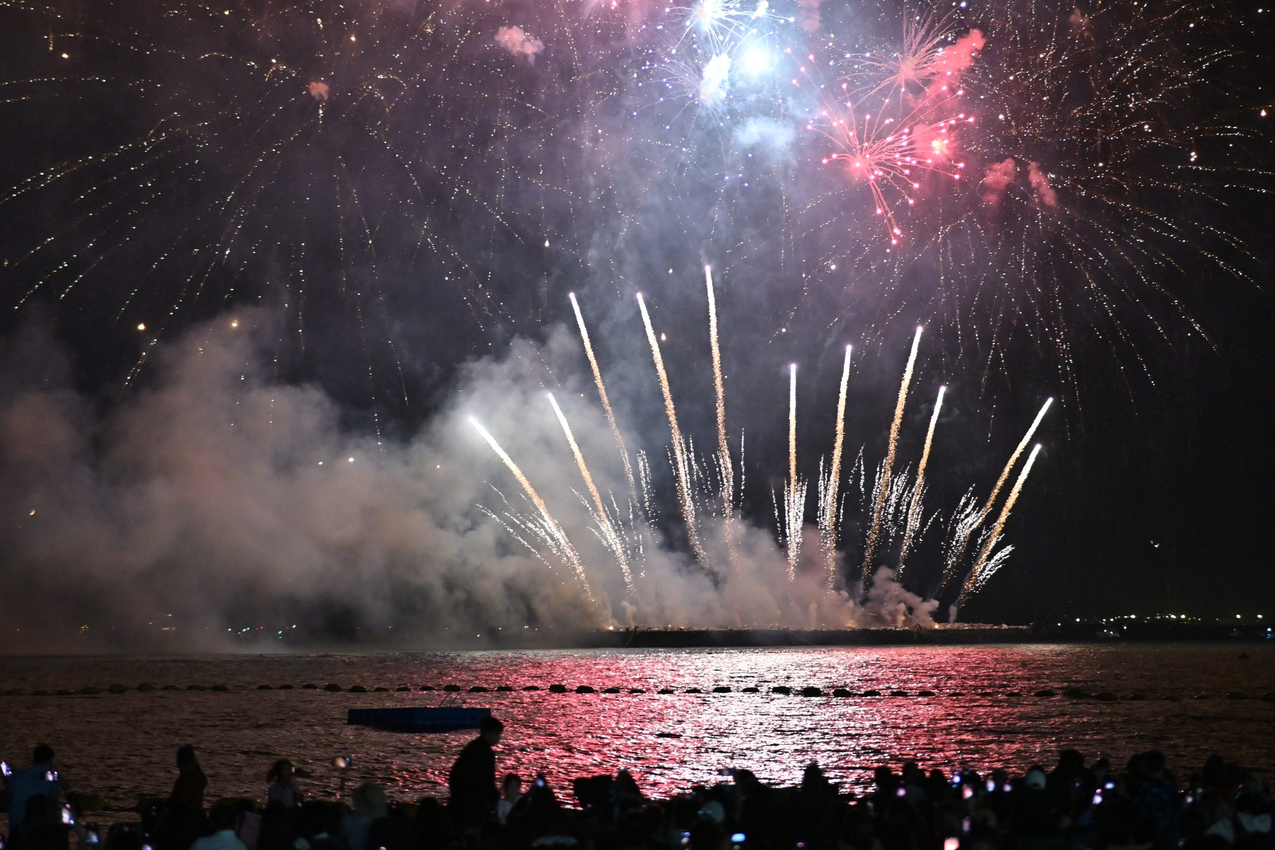 Colourful New Year fireworks bursting over calm water as a crowd watches from the shore