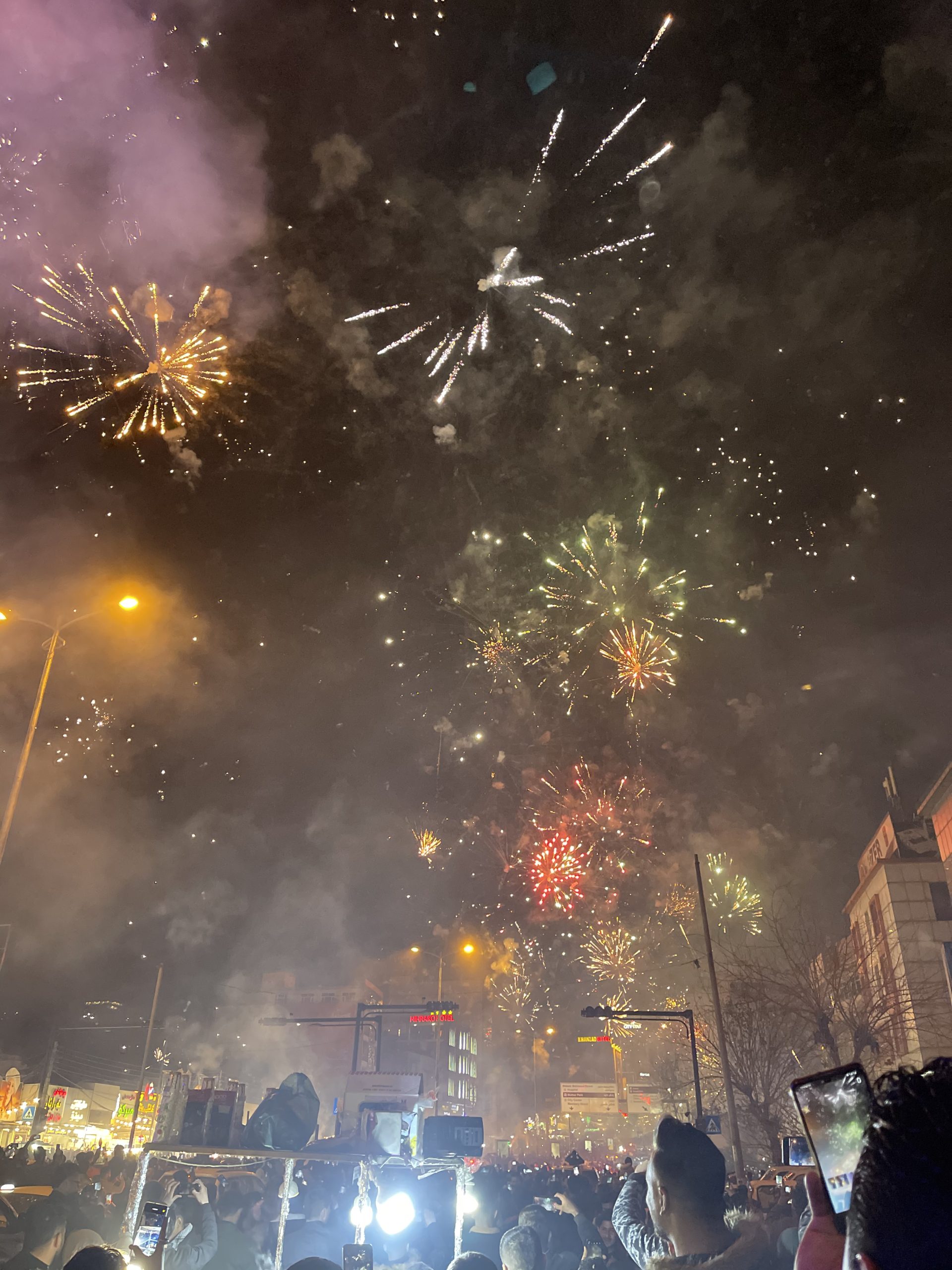 Vertical New Year fireworks filling the sky above a city street packed with people filming